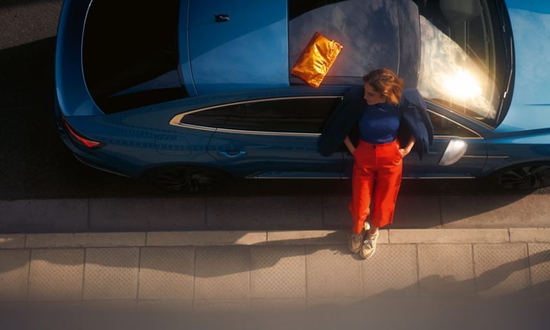 Draufsicht von blauem VW Arteon mit optionalen Ausstattungen Panoramadach und abgedunkelten Scheiben. Eine Frau lehnt an der Tür.