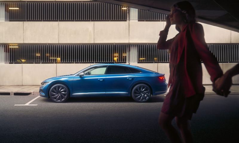 Niebieski Volkswagen Arteon zaparkowany przed parkingiem wielopoziomowym, widok z boku. Na pierwszym planie kobieta w sukience i żakiecie.