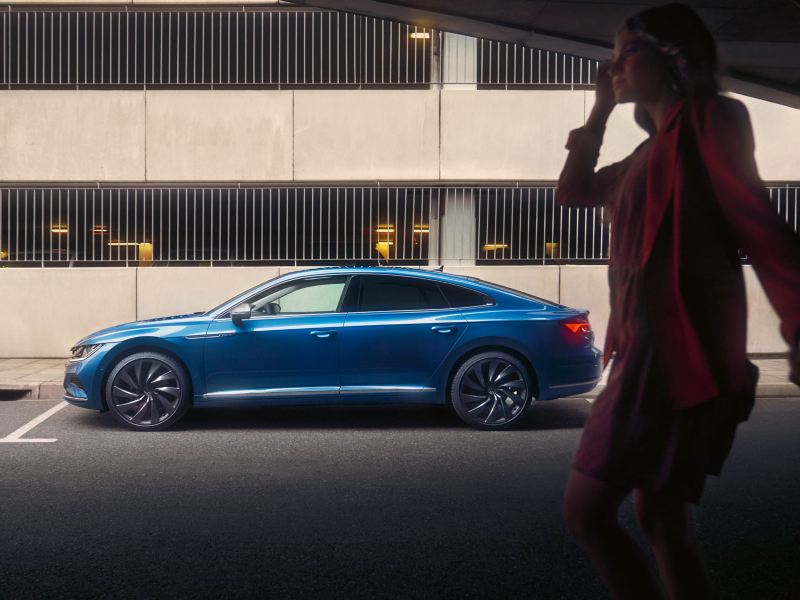 Blauer VW Arteon parkt seitlich vor einem Parkhaus, im Vordergrund eine Frau in Kleid und Jacket.