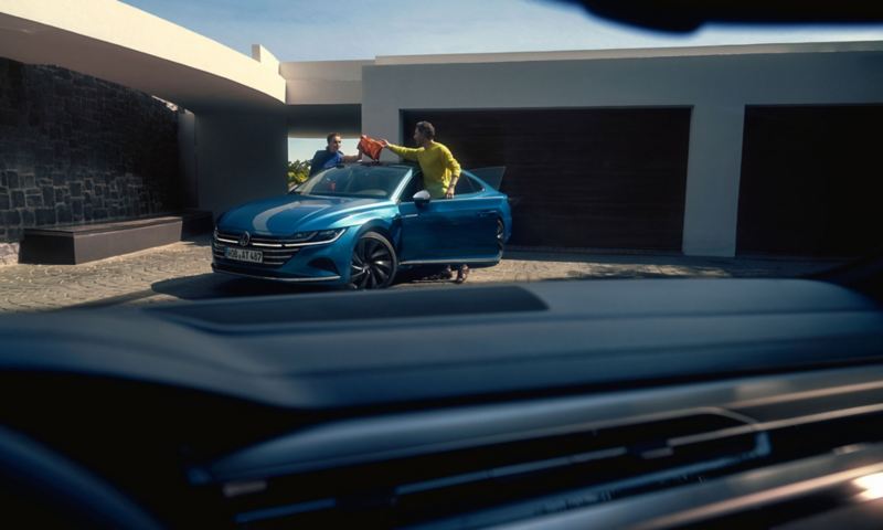 Widok przez przednią szybę samochodu na niebieskiego Arteona przed nowoczesnym budynkiem, kobieta i mężczyzna wsiadają.