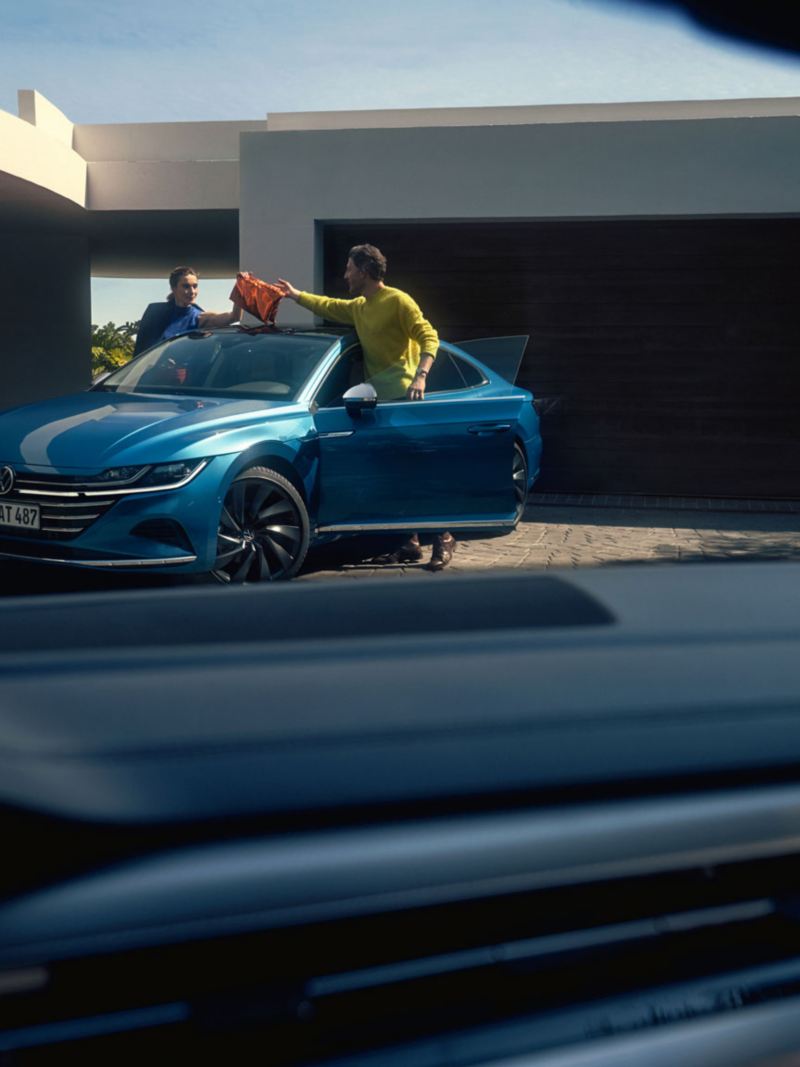 Vue à travers le pare-brise d'une VW Arteon bleue garée devant un immeuble résidentiel moderne, avec un couple qui vient d'arriver.