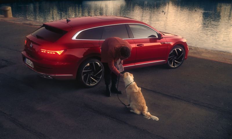 Czerwony Volkswagen Arteon Shooting Brake na parkingu nad wodą, przed nim mężczyzna pochyla się nad psem.