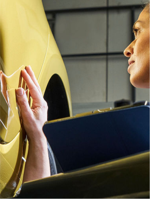 A woman in overalls checks a yellow VW car while holding a clipboard.