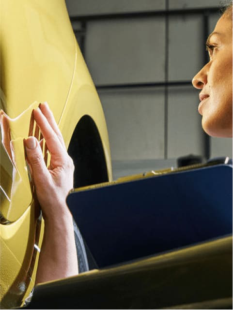 A woman in overalls checks a yellow VW car while holding a clipboard.