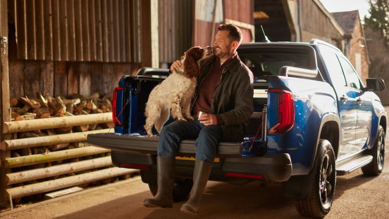 Photo of a man and a dog sitting in the rear section of an Amarok parked next to a barn. 