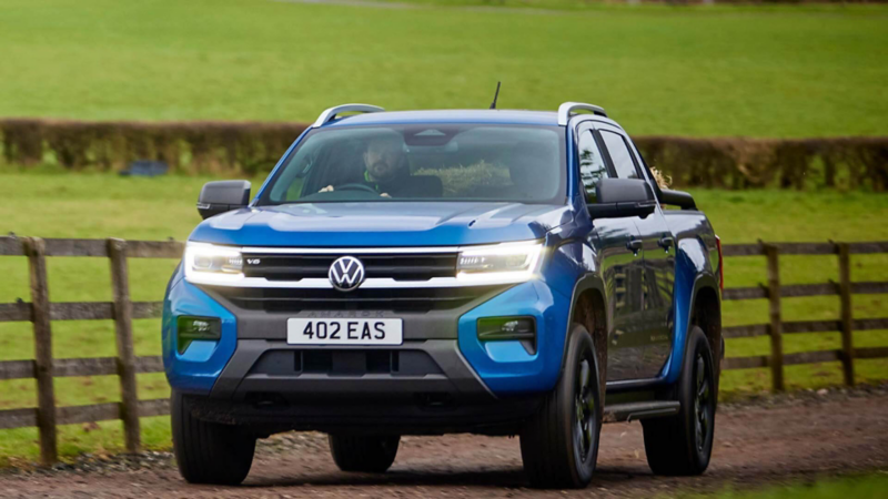 A blue Volkswagen Amarok V6 pickup truck driving on a rural dirt road beside a wooden fence, with green fields in the background. The vehicle's headlights are on, and a bearded driver wearing a green jacket is visible through the windshield. 