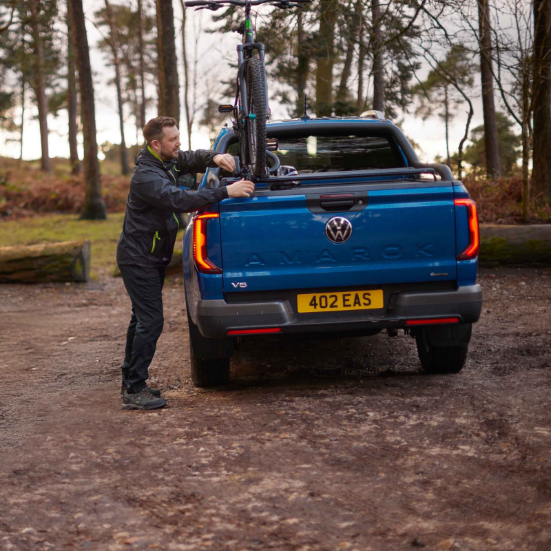 Person securing a bicycle to the back of a Volkswagen Amarok pickup in a wooded area.