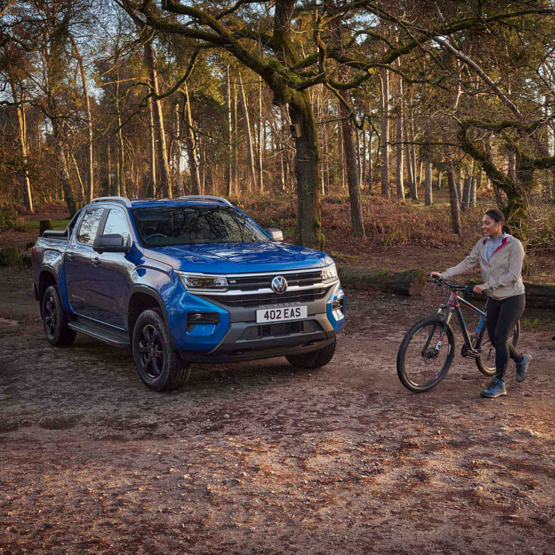 A VW Amarok parked in a woodland area with a woman holding a bike standing next to it.