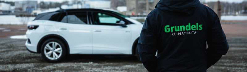 En person med svart jacka med texttryck Gundels klimatruta i framgrunden, vit Volkswagen ID.4 i bakgrunden