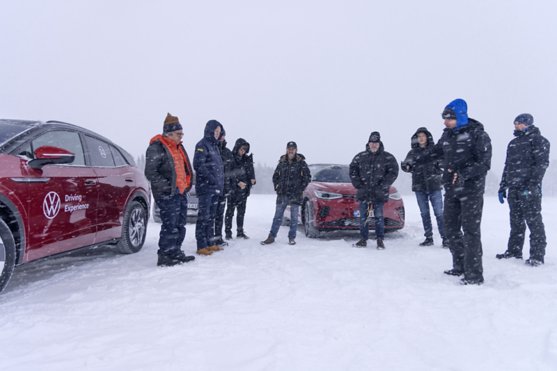 En grupp människor står utomhus på ett snötäckt område under pågående snöfall. Tre elbilar skymtas i bakgrunden.