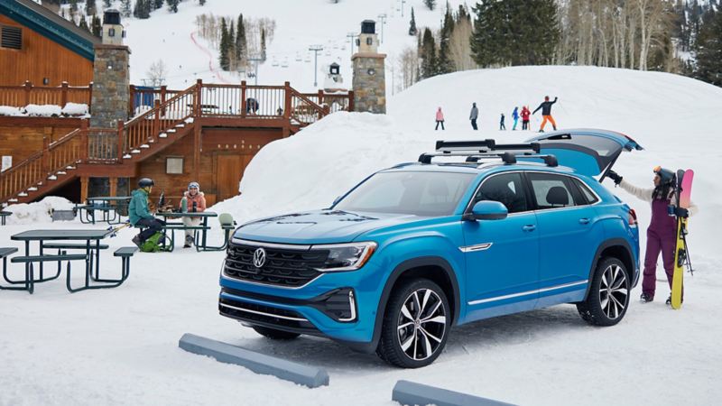 A woman taking snowboard from the trunk of the blue 2024 Volkswagen Cross Sport parked near a snowhill