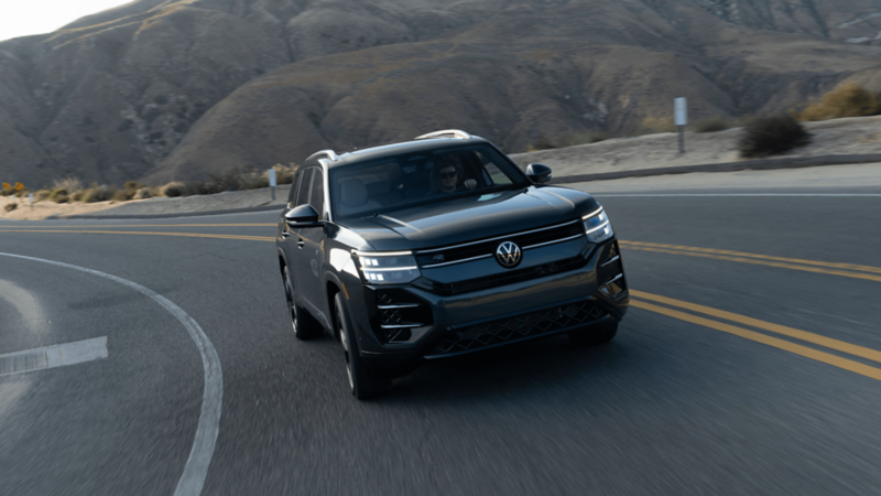 Photo dynamique trois quarts avant d'un VUS Volkswagen Atlas gris foncé négociant un virage sur une route de montagne avec des collines escarpées en arrière-plan et phares éclairés.
