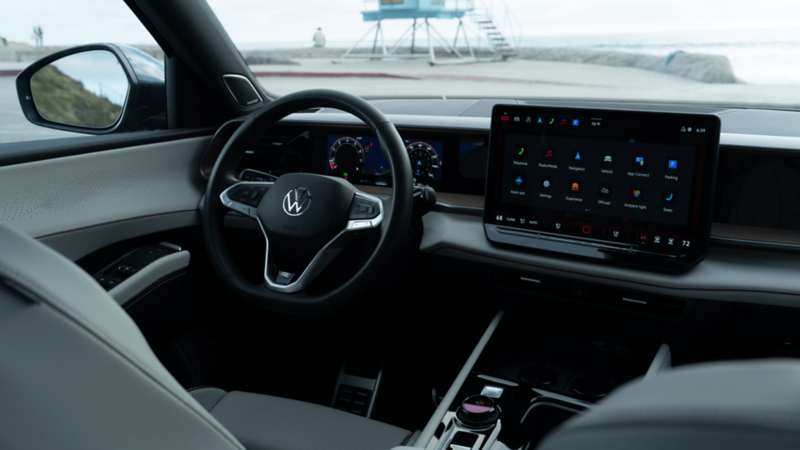 Vue intérieure depuis le siège conducteur montrant le volant gainé de cuir, le combiné d'instruments numérique, l'écran tactile de 15 pouces et la console centrale, avec une plage visible à travers le pare-brise.  