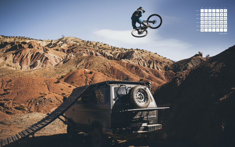 Mountainbiker laver et hop i luften fra en rampe bygget på en campervan i et tørt, rødt bjerglandskab, med en lille august 2026-kalender i billedets hjørne.