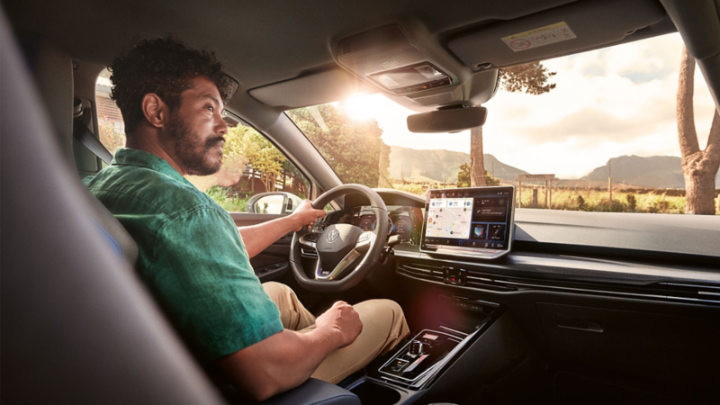 Une personne au volant d'une Volkswagen équipée d'un tableau de bord numérique et d'un écran de navigation, roulant sur une route avec un paysage et des montagnes en arrière-plan.