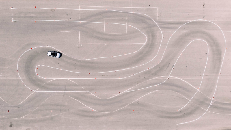 Vue aérienne d'une voiture blanche naviguant sur un parcours d'autocross sinueux marqué de lignes blanches et de petits cônes orange sur une surface asphaltée.
