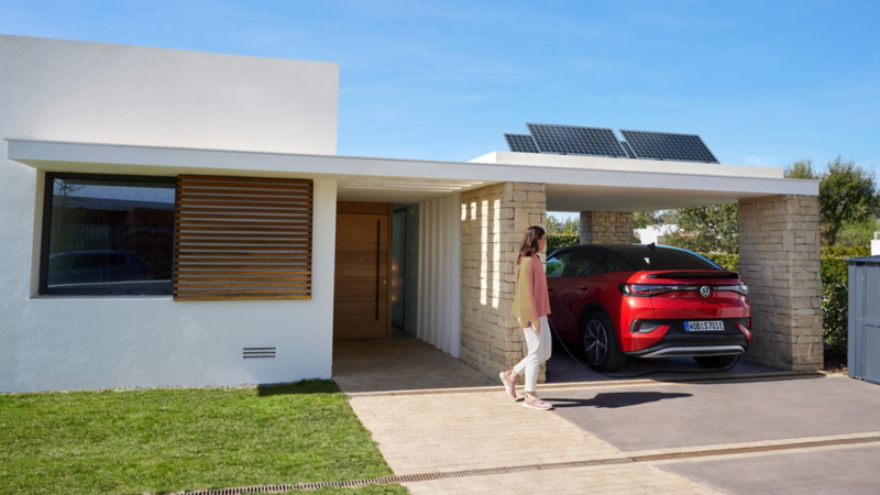 Ein  ID.5 GTX steht unter einem modernen Carport neben einem weißen, minimalistischen Wohnhaus. Eine Frau geht auf das Fahrzeug zu.