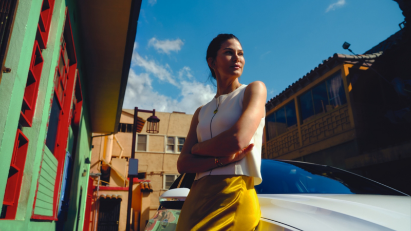 Une femme portant un haut blanc et des pantalons jaunes est appuyée contre une Volkswagen. Elle est entourée d’immeubles colorés, sous un ciel bleu.