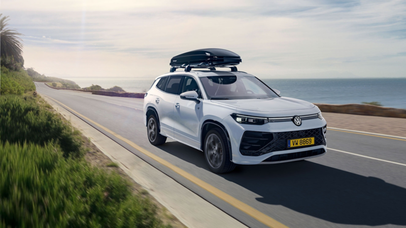 Une VW Tayron blanche avec un coffre de toit noir roule sur une route qui longe la mer.