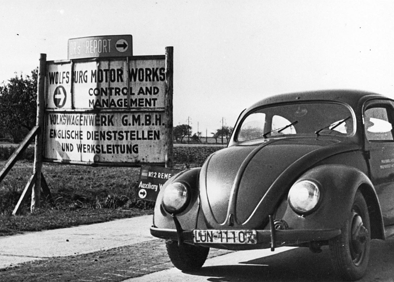 Historic black-and-white photo of a Volkswagen Beetle parked near Wolfsburg Motor Works sign marking early factory site.