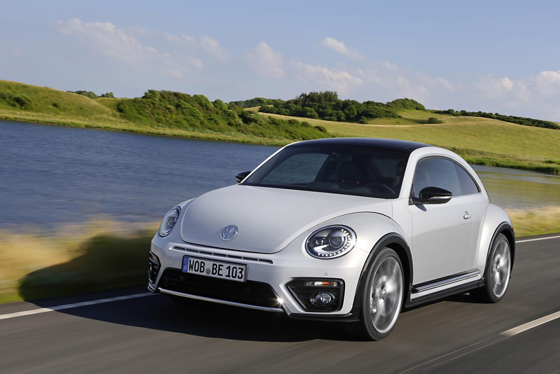 White Volkswagen Beetle driving along a lakeside road, shown from the front three‑quarter angle