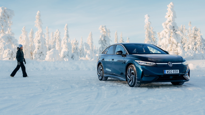 En blå ID.7 står parkerad i ett vinterlandskap, en vinterklädd person gåt mot bilen