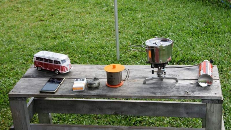 車主露營的桌面擺設，木製露營桌上擺有紅白雙色California 模型車、簡易水煮壺、鋼杯、手機和菸包