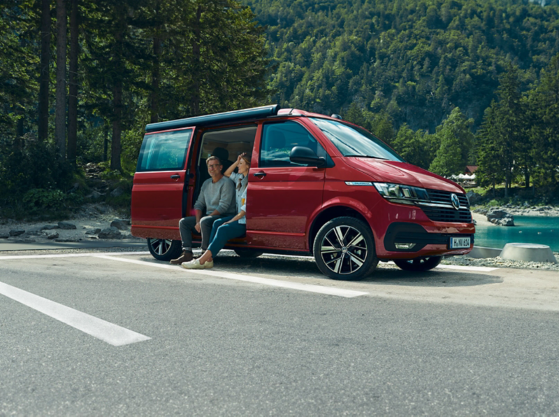 un VW California 6.1 est garée au bord d’un lac près d'une forêt avec 2 personnes assis sur le côté