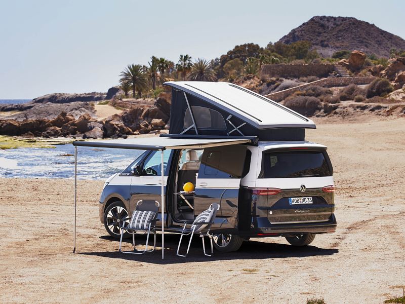 Ein VW California mit hochgestelltem Aufstelldach und ausgefahrener Markise und Faltstühlen am Strand stehend.