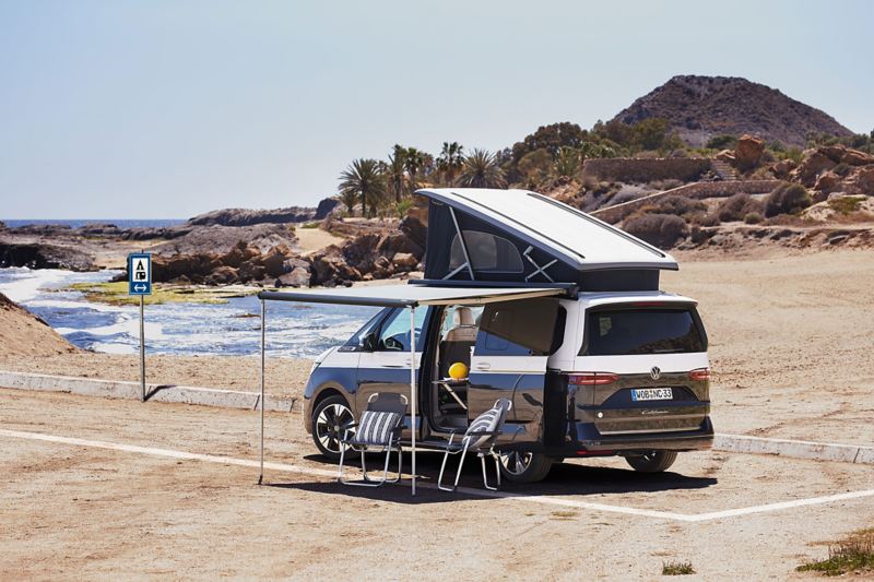 Une VW California avec le toit relevable, l'auvent étendu et les chaises pliantes posées sur la plage.