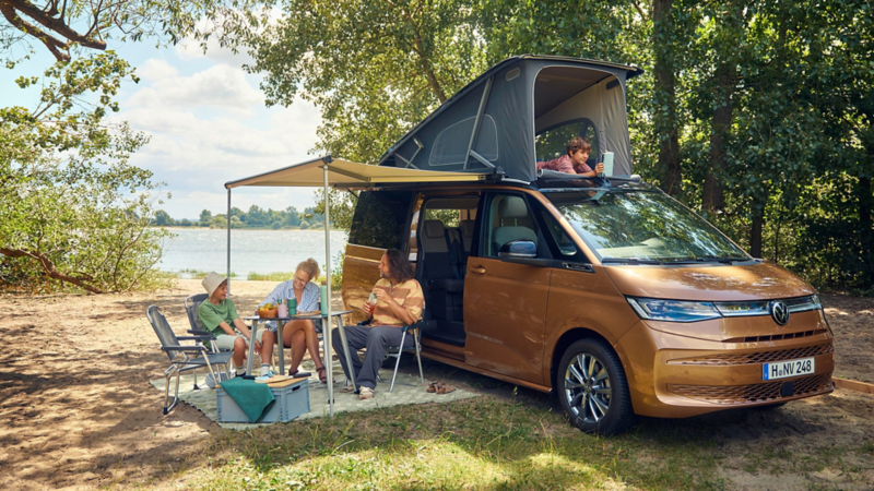 California mit geöffnetem Aufstelldach auf einem Campingplatz, davor mehrere Personen