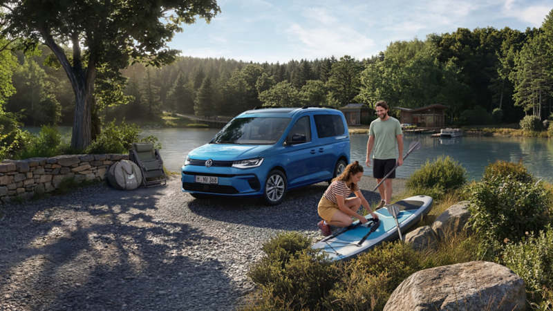 Caddy California an einem See, daneben zwei Personen mit SUP-Board