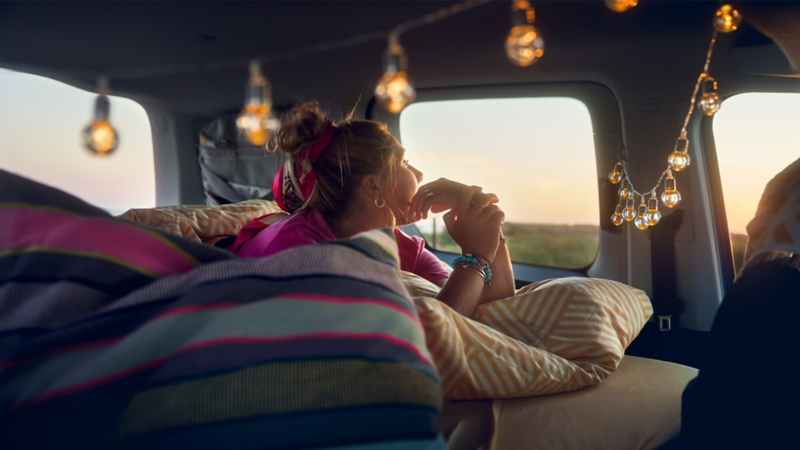 Eine Frau liegt in ihrem VW Camper, der innen mit einer Girlande aus Leuchten dekoriert ist.