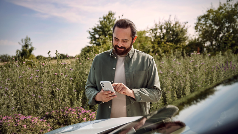 Ein Mann steht in einer grünen Landschaft neben einem VW ID. und bedient sein Smartphone.
