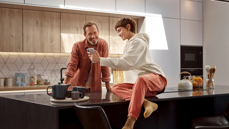   Far og datter kigger i køkkenet på en mobiltelefon, der viser Volkswagen appen.