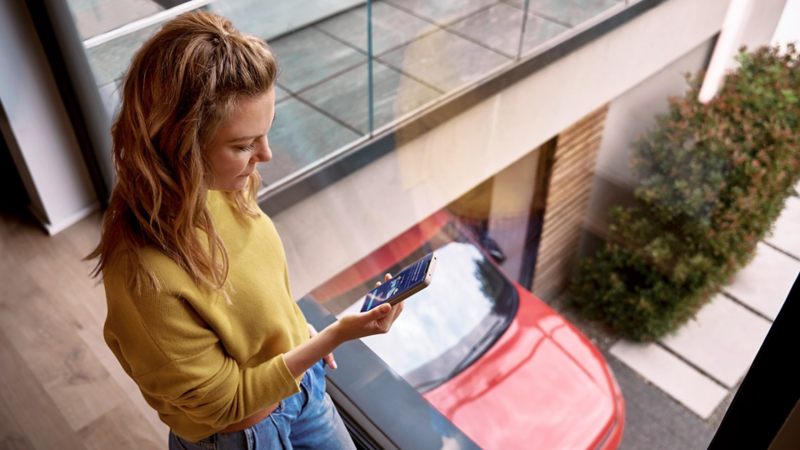 Frau schaut auf Handy. Im Hintergrund ein roter Volkswagen.  In der rechten oberen Ecke die Logos von App Store und Google Play Store.