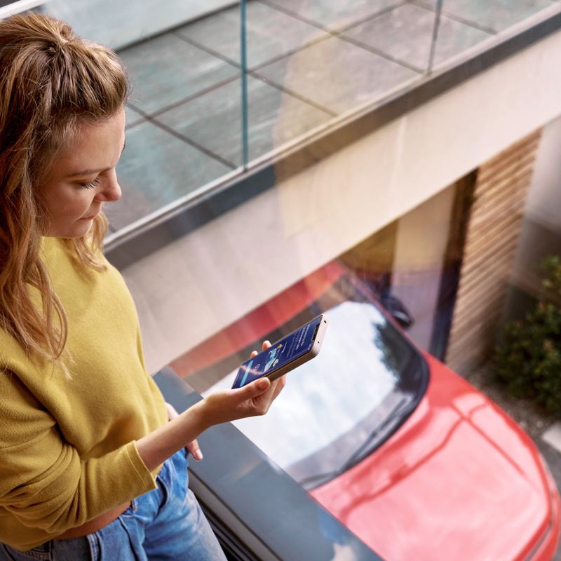 Frau schaut auf Handy. Im Hintergrund ein roter Volkswagen.