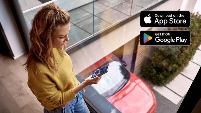 Une femme regarde son téléphone mobile. En arrière-plan, une Volkswagen rouge. Sur le coin supérieur droit, les logos de Apple Store et Google Play Store sont visibles.