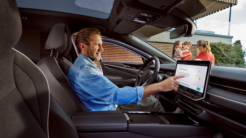 Mann sitzt in seinem Volkswagen und berührt das Display, im Hintergrund seine Familie