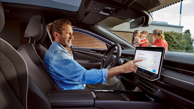 Mann sitzt in seinem VW ID.7 und berührt das Display, im Hintergrund eine Frau und zwei Kinder