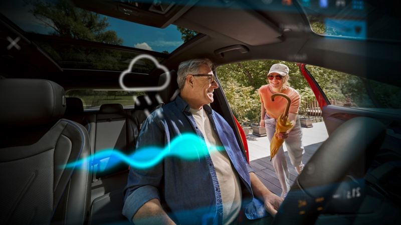 Un homme âgé est assis dans sa Volkswagen, une femme lui tend un parapluie.