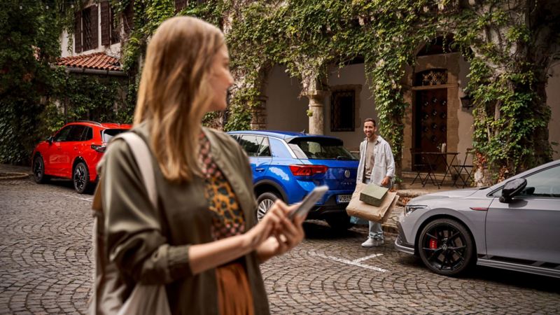 Une femme en avant-plan avec téléphone mobile, un homme avec des sacs entre deux voitures garées