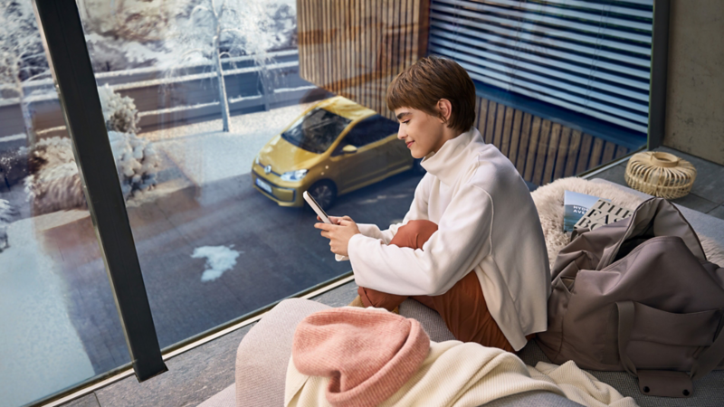 Persona dentro de una casa revisando su teléfono inteligente, con un automóvil Volkswagen amarillo visible a través de la ventana, lo que resalta su elegante propiedad.