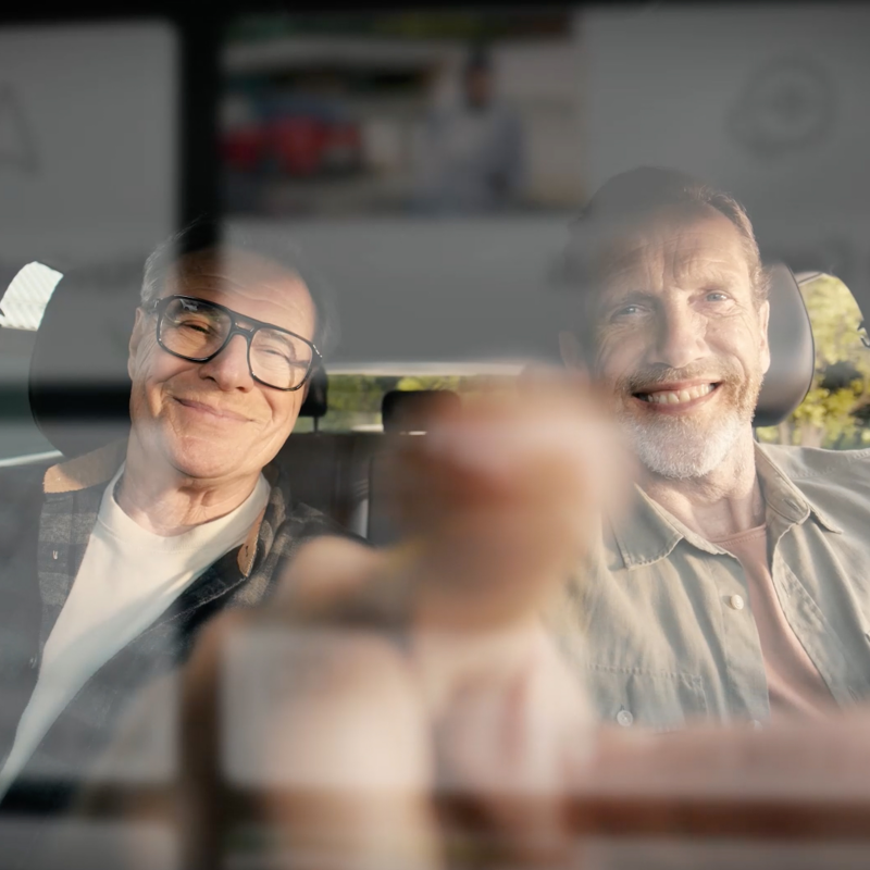 Deux hommes sont assis dans une voiture, photographiés de l’extérieur à travers le pare-brise.