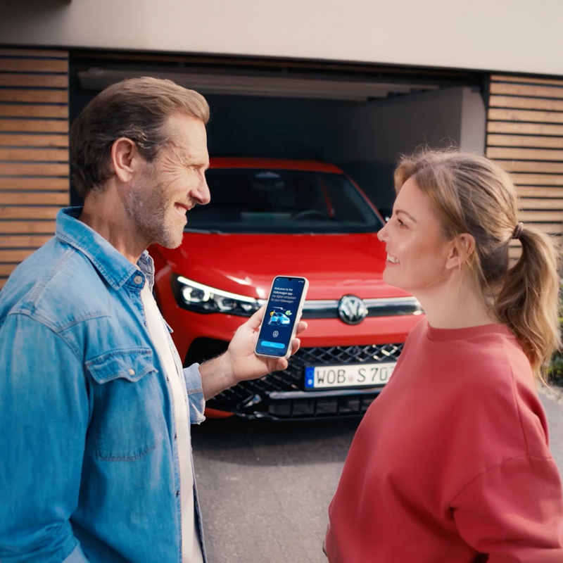 A woman and a man holding a smartphone standing in front of a Volkswagen, switching between looking at each other and the vehicle.