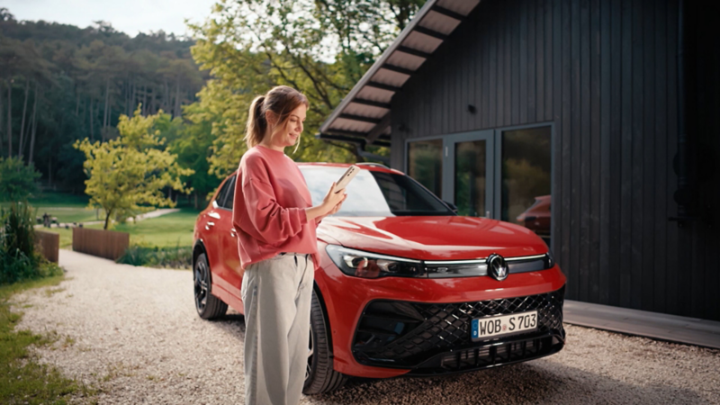 Une femme, smartphone à la main, se tient debout devant sa voiture VW rouge. 