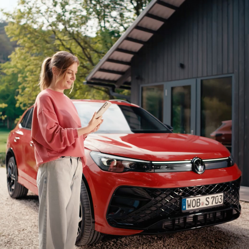 Frau steht vor ihrem Volkswagen und schaut auf ihr Handy.