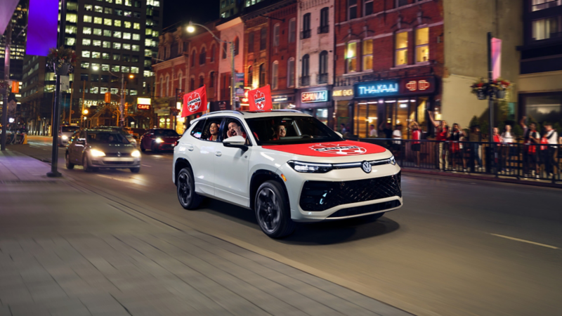 Tiguan Volkswagen blanc avec drapeaux de Canada Soccer roule en centre‑ville le soir; des passants acclament sur le trottoir.