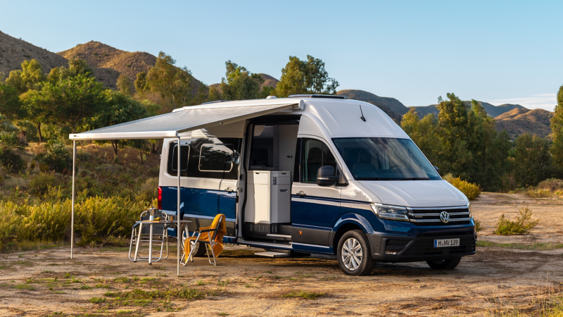 VW Grand California mit aufgestellter Markise parkt in der Natur.