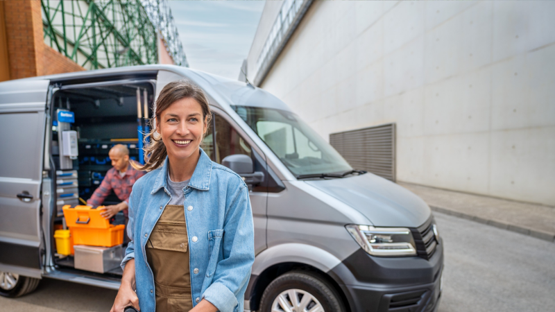 Eine Handwerkerin vor VW Crafter mit geöffneter Schiebetür, welcher auf einem Industriegelände parkt